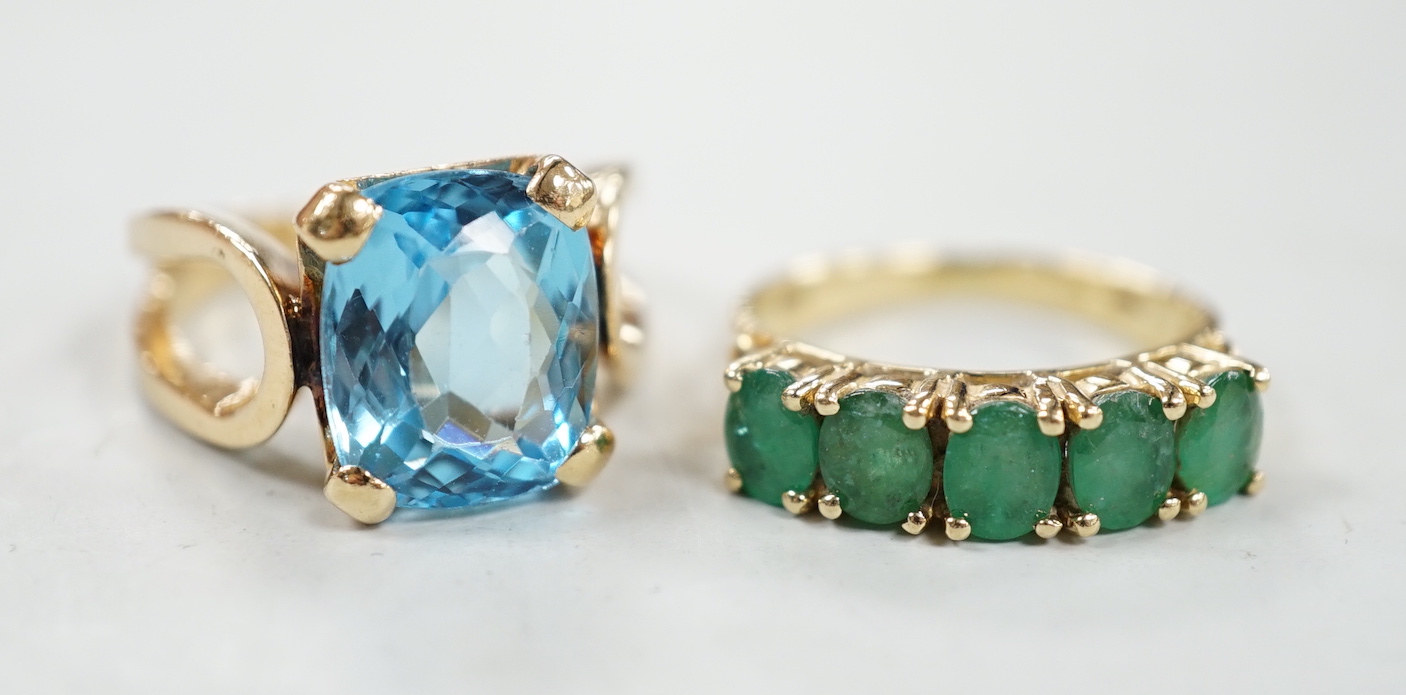 A modern 14k yellow metal and cushion cut blue topaz set dress ring, size K/L and a 14k yellow metal and five stone emerald set half hoop ring, size L/M, gross weight 10.5 grams.